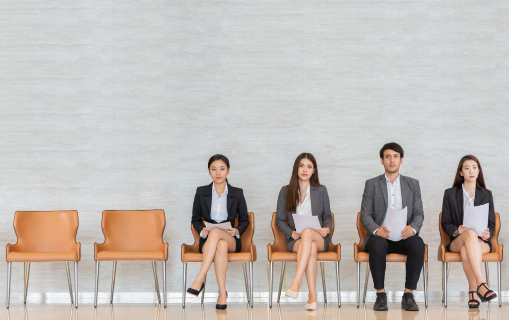 Quatro pessoas, três mulheres e um homem, aguardam entrevista de emprego em cadeiras bege.