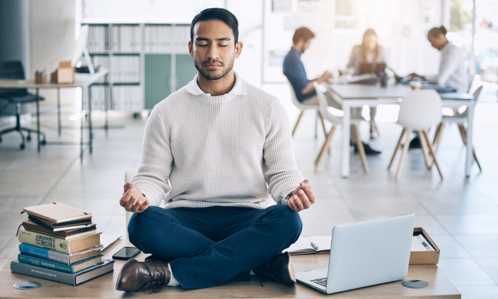 Homem em ambiente de trabalho, sentado no chão em posição de lótus fazendo meditação com laptop aberto ao seu lado.