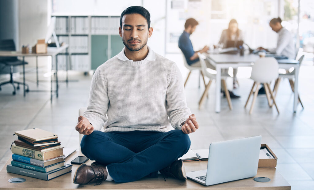 Homem em ambiente de trabalho, sentado no chão em posição de lótus fazendo meditação com laptop aberto ao seu lado.