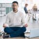 Homem em ambiente de trabalho, sentado no chão em posição de lótus fazendo meditação com laptop aberto ao seu lado.