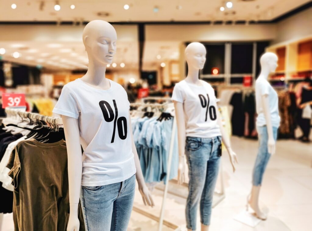 Dois manequins femininos com camiseta branca e símbolo de porcentagem e calça jeans, em fundo de loja de roupas.