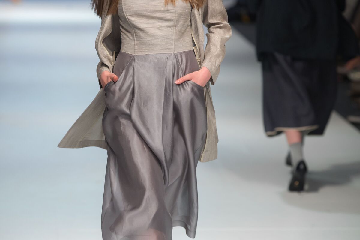 Modelo com rosto cortado da imagem desfila com mãos nos bolsos de vestido cinza-roxo-prateado, representando tendências da moda