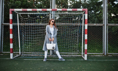 Mulher com casaco cinza e bolsa grande em frente a gol de futebol