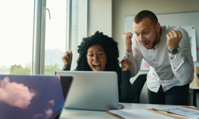 Homem branco de pé e mulher negra sentada comemoram frente a um computador, remetendo a promoção