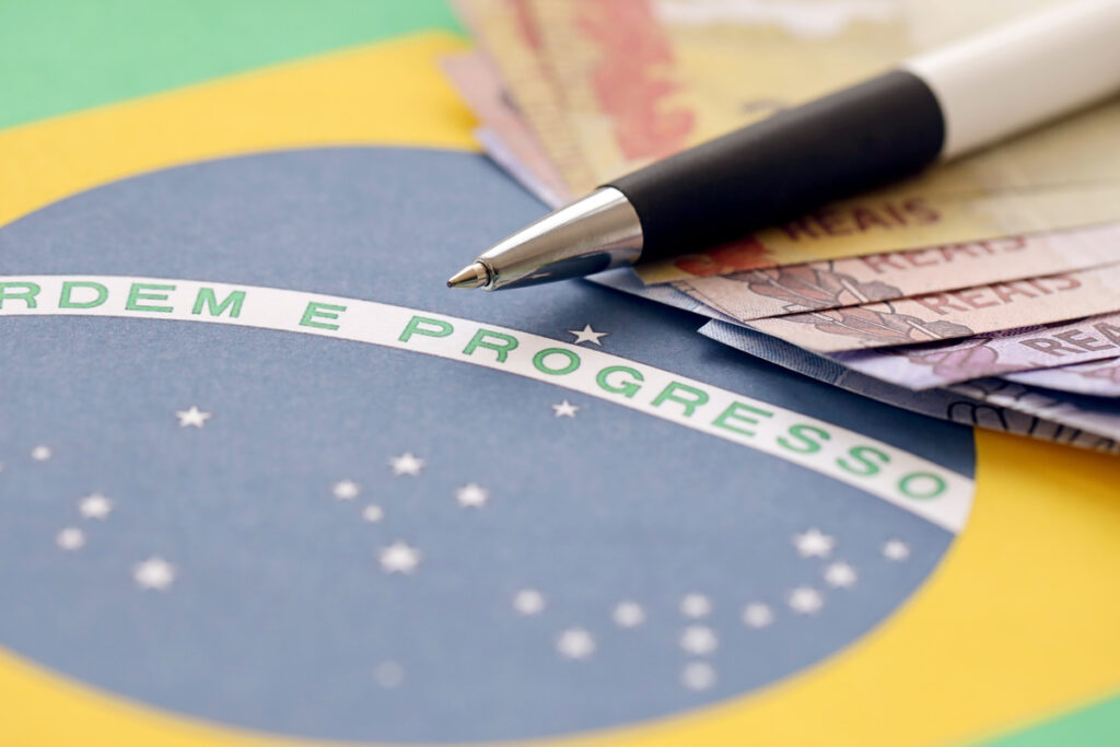 Close em bandeira do Brasil com cédulas de dinheiro em reais, e caneta por cima.
