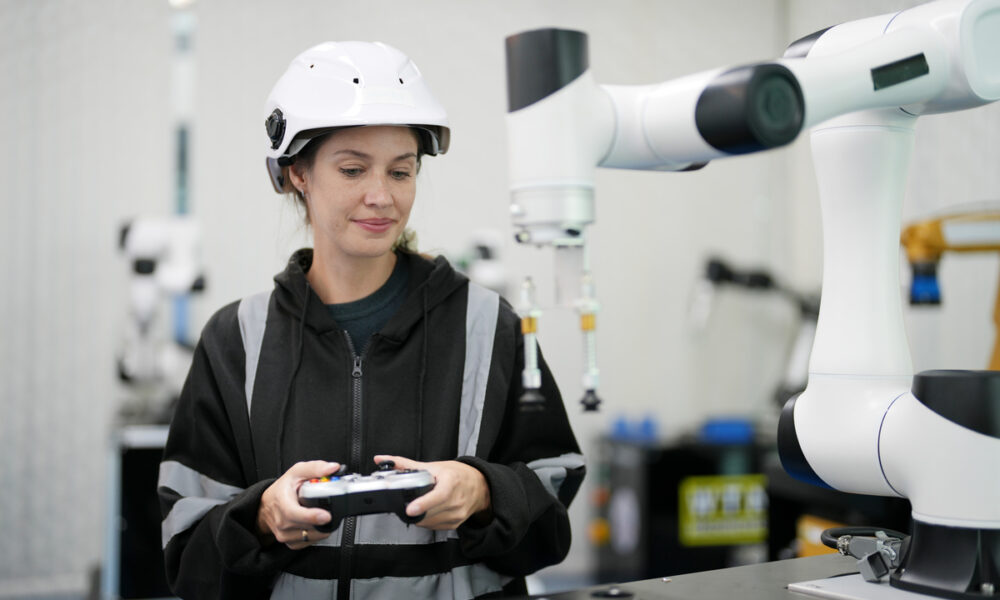 Mulher com roupa de proteção e capacete industrial com controle na mão comandando braço mecânico