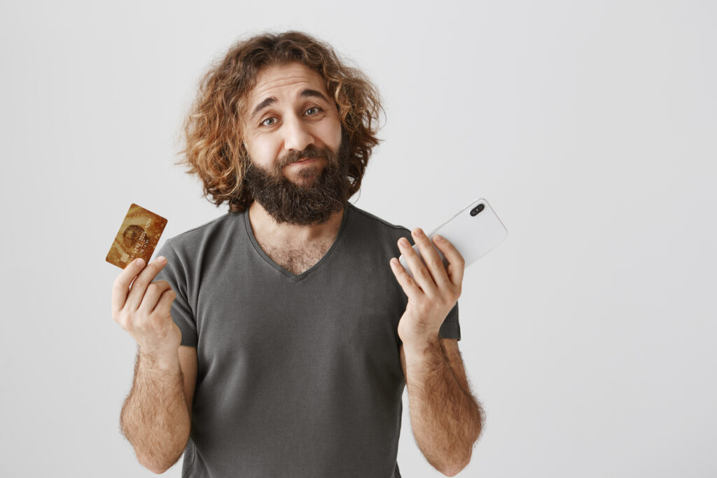 Retrato de um homem triste e arrependido com barba franzindo a testa e encolhendo os ombros, mostrando cartão de crédito e smartphone