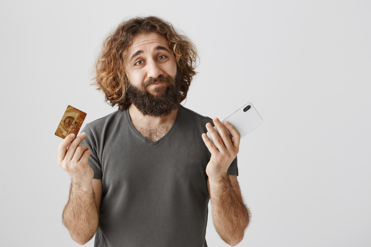Retrato de um homem triste e arrependido com barba franzindo a testa e encolhendo os ombros, mostrando cartão de crédito e smartphone; rotativo