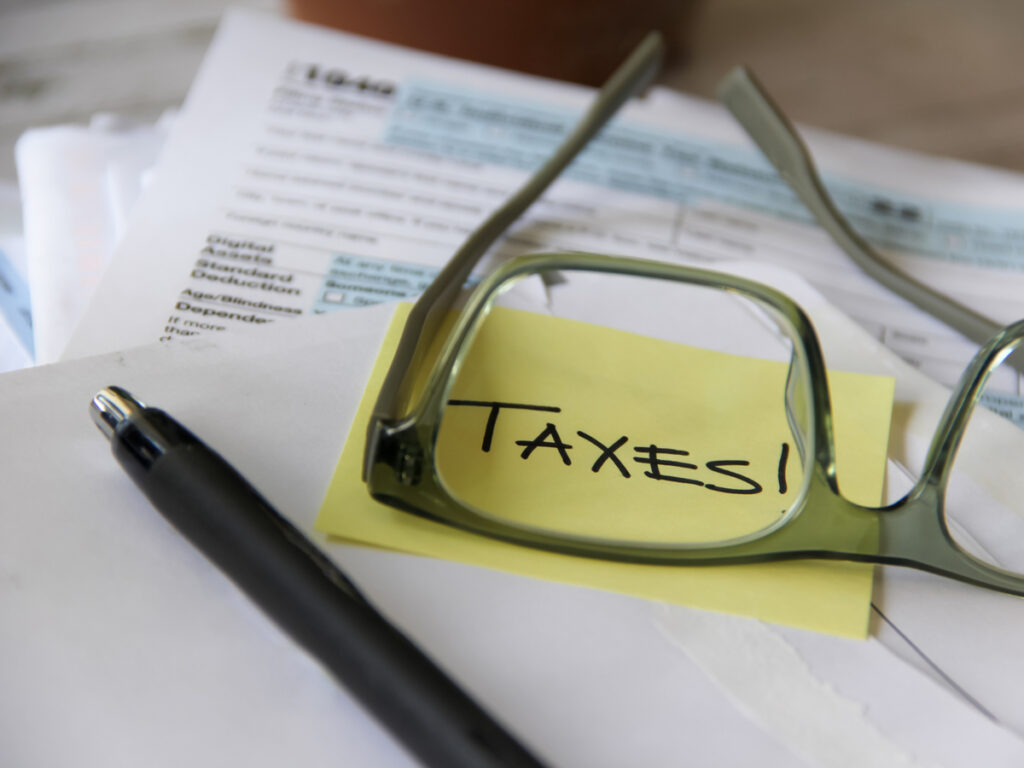 Papel com post-it amarelo escrito "Taxes" sendo ampliado por óculos e lápis perto