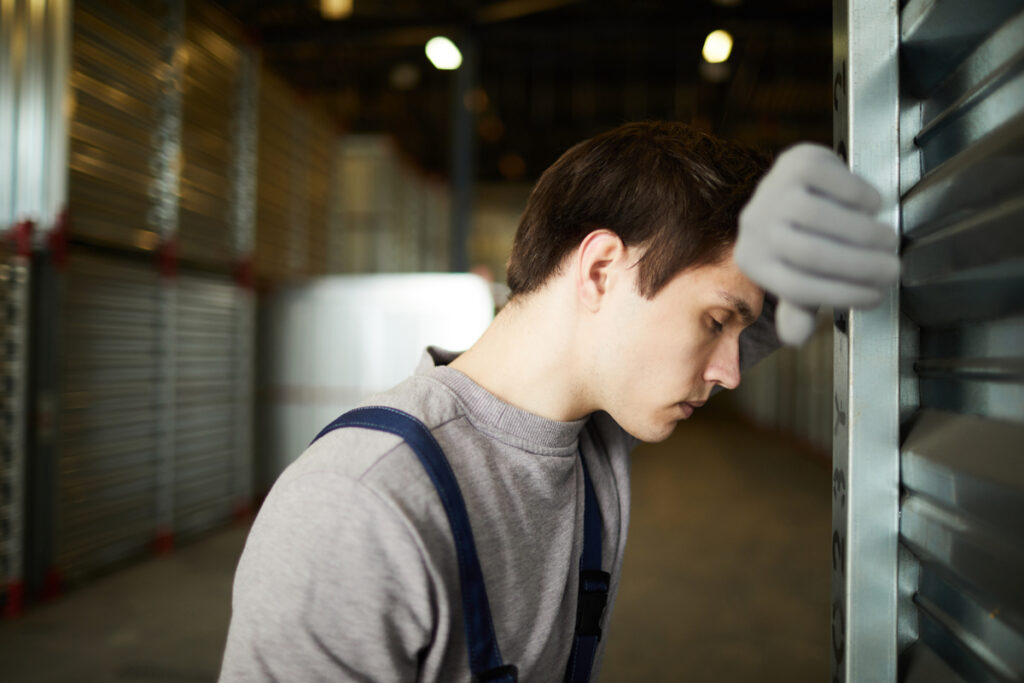 Trabalhador branco com camiseta de mangas cinza, jardineira e luva se apoia em parede, exasperado por saúde mental no trabalho ruim