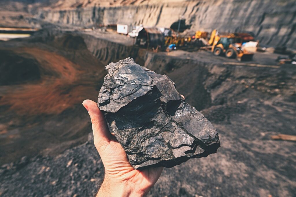 Estação de mineração com máquinas trabalhando, e, em primeiro plano, mão de pessoa segurando pedra preta de minério de ferro, como produzido pela Vale