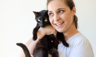 Mulher de blusa branca e olhos verdes abraçando gato preto e sorrindo