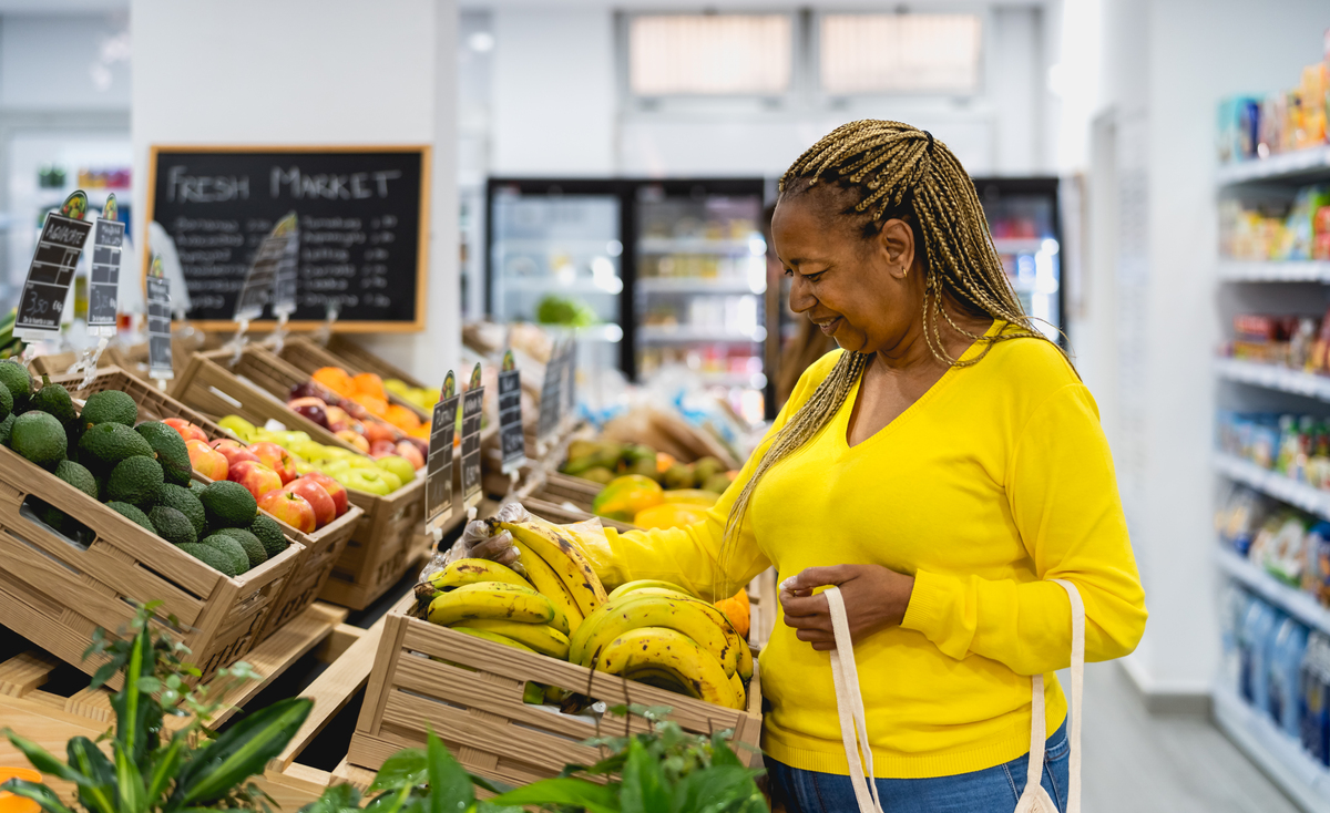 Mulher negra de blusa amarela pega bananas no supermercado, sorrindo, com sacola na mão