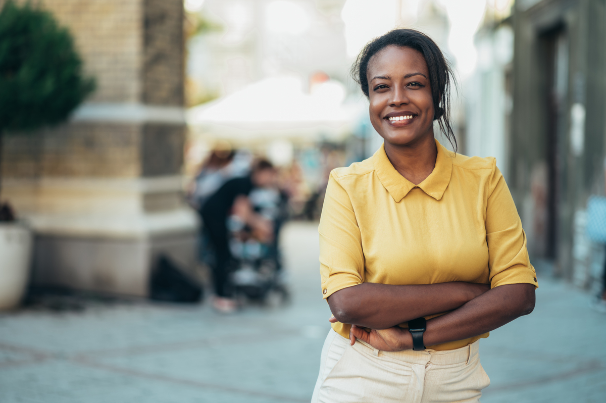 Mulher negra na calçada com braços cruzados e sorrindo para a câmera.