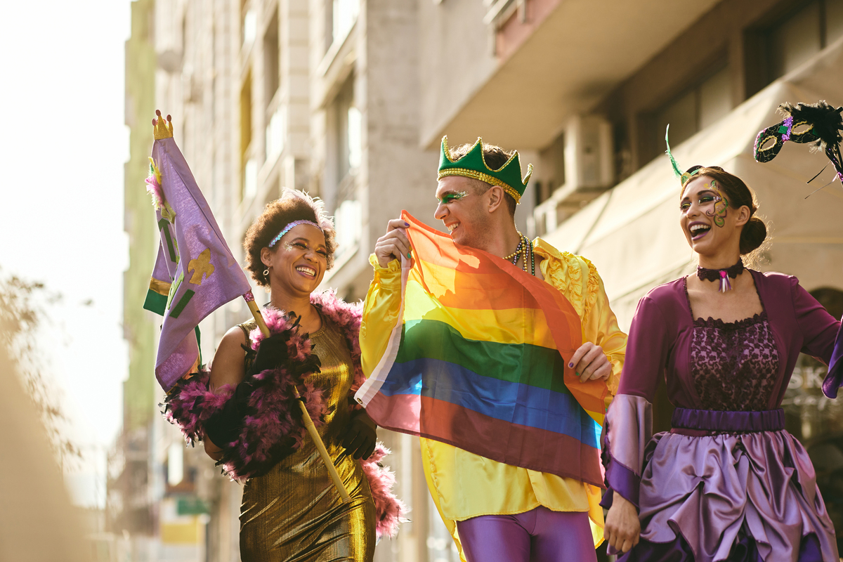 Grupo de três jovens fantasiados para o carnaval. Da esquerda para a direita são uma mulher negra com vestido dourado e segurando uma bandeira roxa; um homem com coroa verde segurando uma bandeira lgbt na frente do corpo e uma mulher com roupa roxa de princesa.