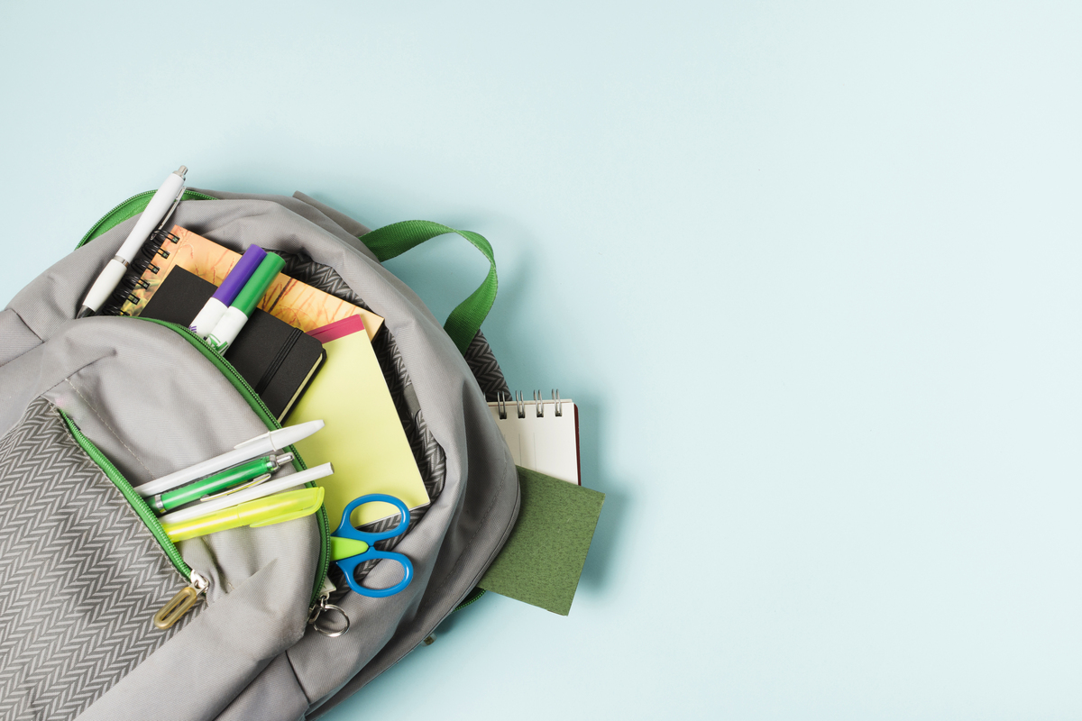 Fundo azul claro, com mochila aberta no canto esquerdo e materiais escolares como caneta, régua e tesoura saem para fora.