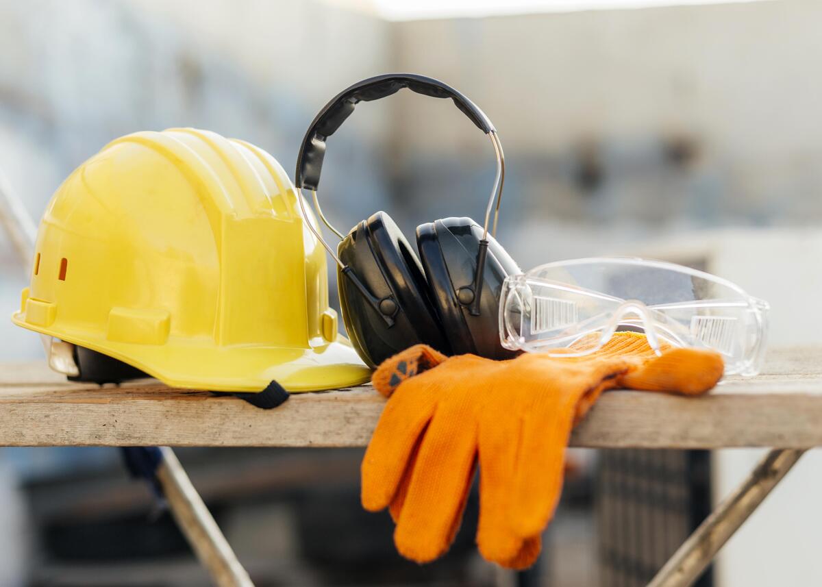 Equipamentos de segurança de trabalho: capacete amarelo, fones de ouvido para ruído, luvas, óculos transparentes.