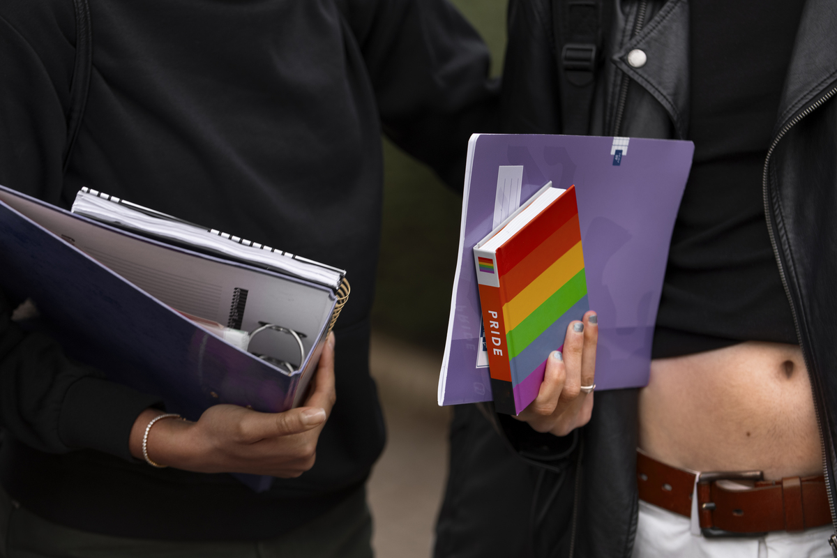 Close em mãos de pessoas em ambiente de trabalho, e uma delas segura livro com cores da bandeira lgbt escrito "pride"