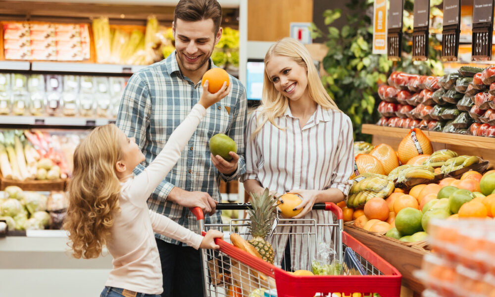 Família jovem feliz em pé com um carrinho e escolhendo frutas no supermercado