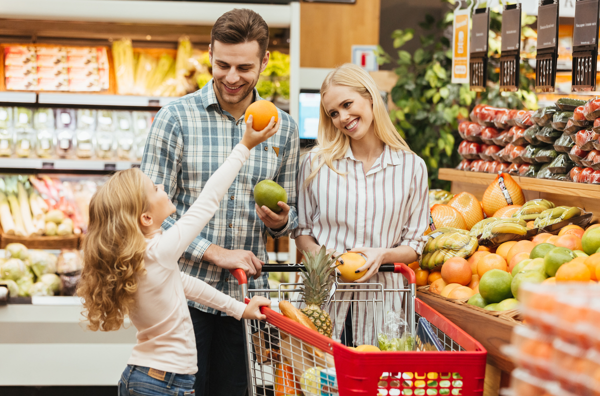 Família jovem feliz em pé com um carrinho e escolhendo frutas no supermercado