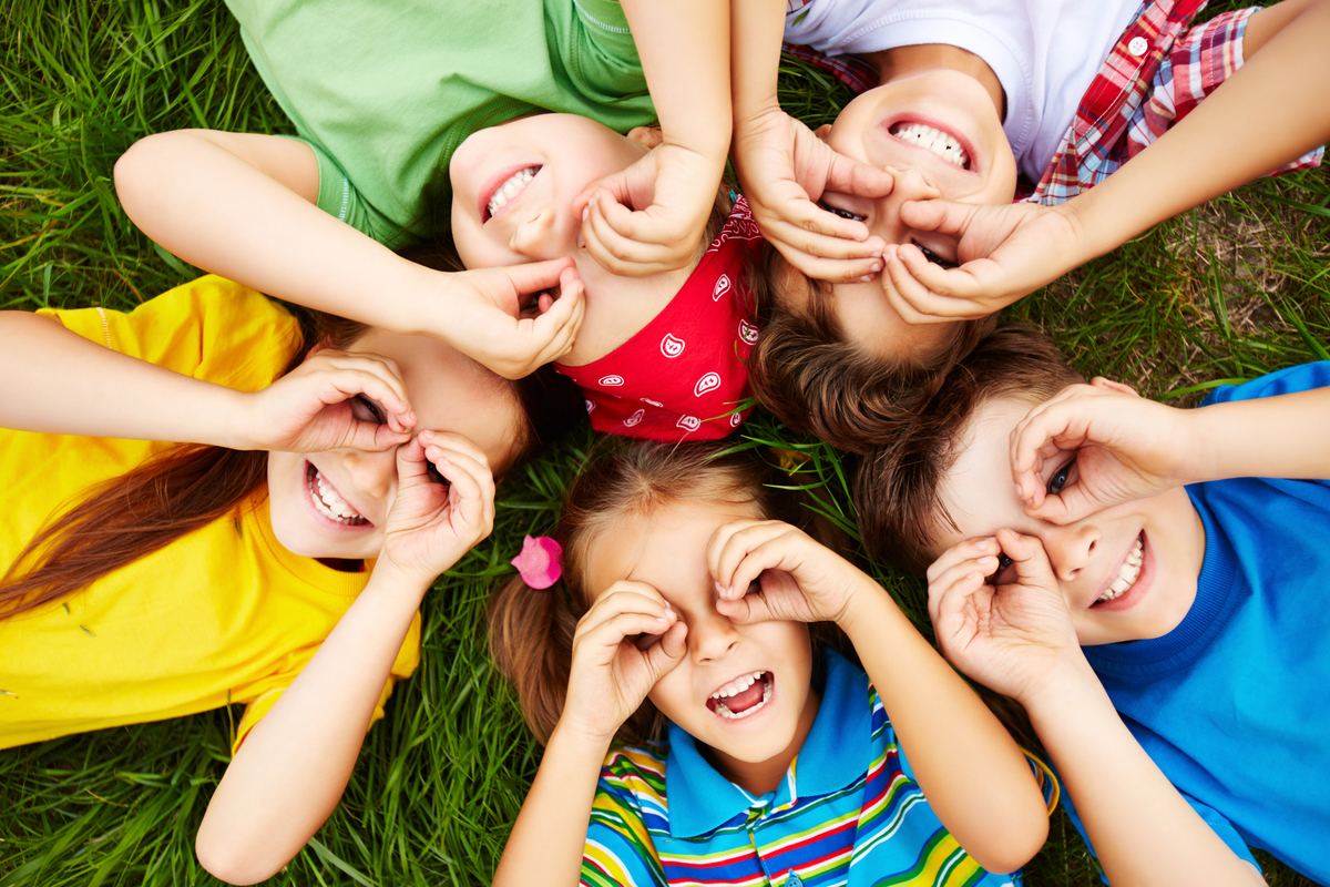 Cinco crianças deitadas na grama, sorrindo e fazendo óculos com as mãos nos olhos.