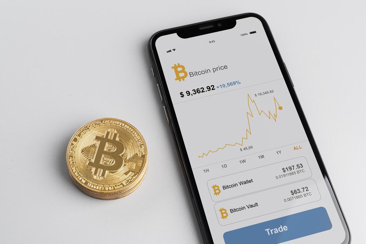 Celular com página de Bitcoin aberta, com moeda com "B" talhado ao lado; criptomoeda