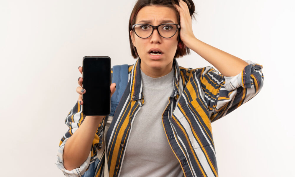 Mulher de óculos com a mão na cabeça e boa aberta mostrando preocupação enquanto mostra o celular com a outra mão.