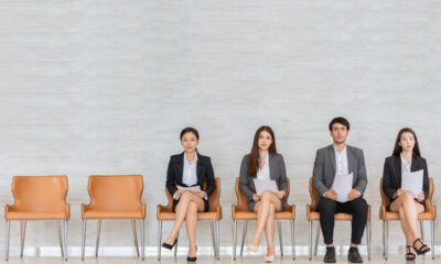 Quatro pessoas, três mulheres e um homem, aguardam entrevista de emprego em cadeiras bege.