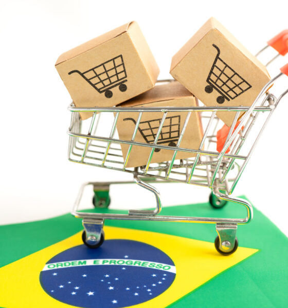 Carrinho virtual de supermercado cheio de caixas de compras, em cima de uma bandeira do Brasil ilustra investimentos no varejo brasileiro.
