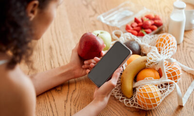 Mulher segura maçã de frente a cesta de frutas com uma mão, e celular com outra.