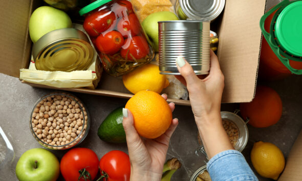 Mão feminina separa alimentos diversos e os coloca em caixa de doação, ilustrando a notícia de que a Connecting Food distribuiu mais de 48 toneladas de alimentos