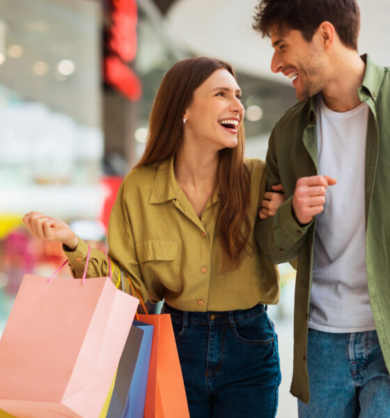 Casal formado por homem e mulher sorri, enquanto olham um para o outro em um shopping, com sacolas na mão.