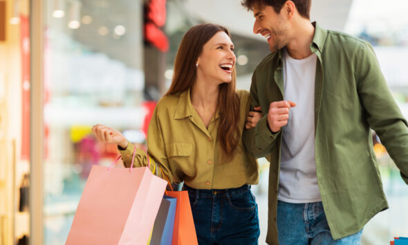 Casal formado por homem e mulher sorri, enquanto olham um para o outro em um shopping, com sacolas na mão.