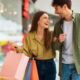 Casal formado por homem e mulher sorri, enquanto olham um para o outro em um shopping, com sacolas na mão.