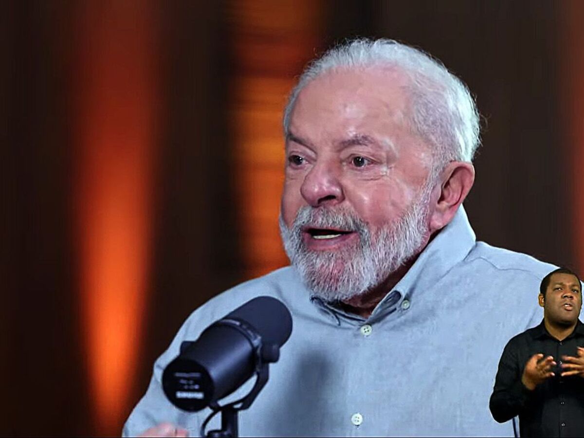 Lula fala em microfone com camisa azul, à frente de fundo vermelho