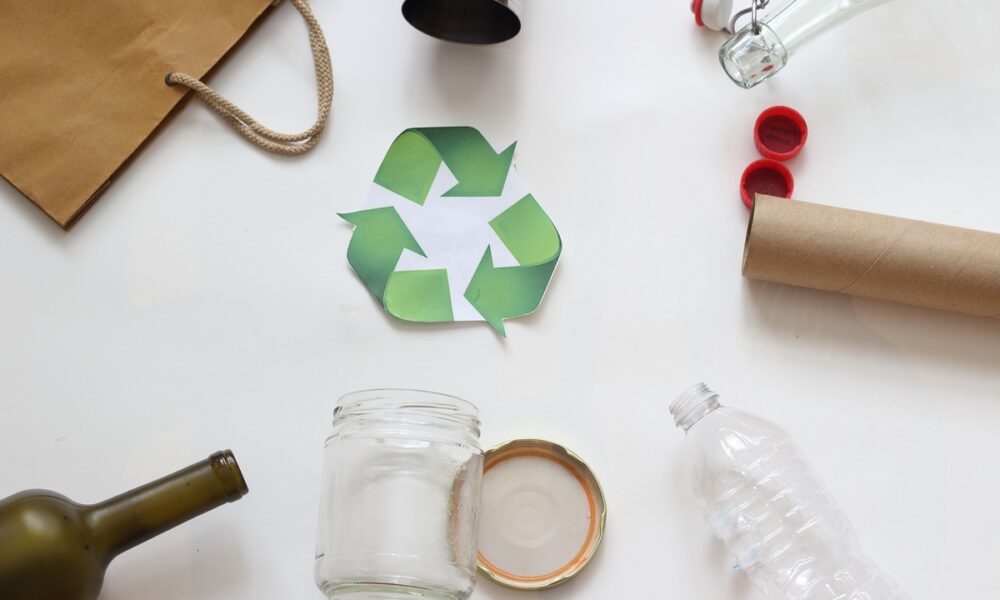 Símbolo verde de reciclagem é cercado de resíduos sólidos como papel, vidro e plástico.