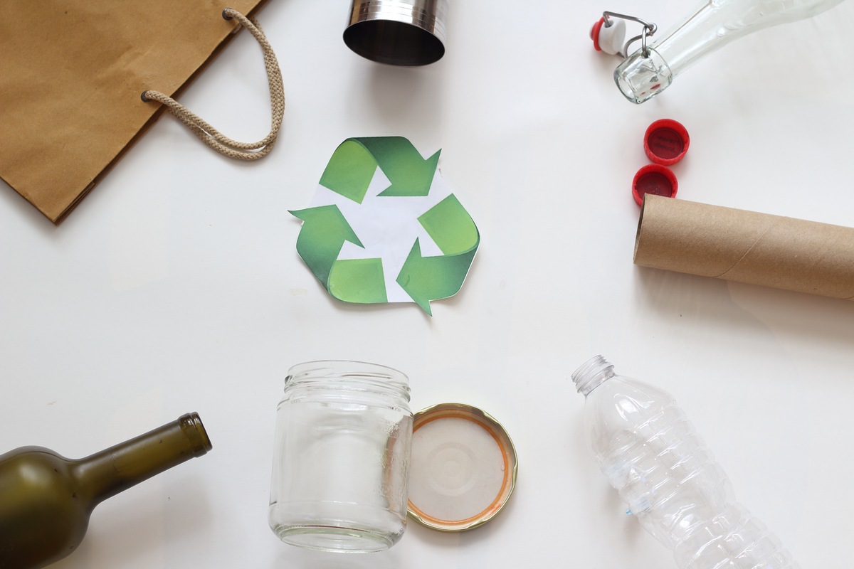 Símbolo verde de reciclagem é cercado de resíduos sólidos como papel, vidro e plástico.