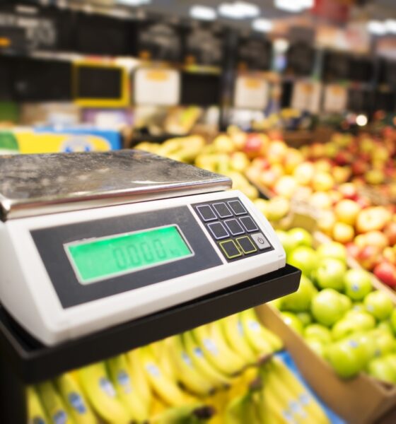 Máquina de pesar alimentos com frutas e legumes ao fundo; varejista