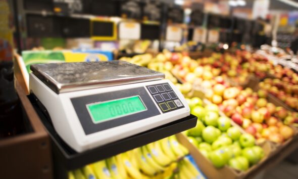 Máquina de pesar alimentos com frutas e legumes ao fundo; varejista