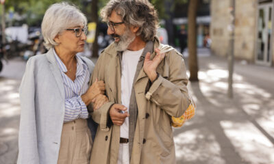 Um casal de idosos segura no braço um do outro andando na rua, enquanto se olham