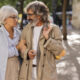Um casal de idosos segura no braço um do outro andando na rua, enquanto se olham