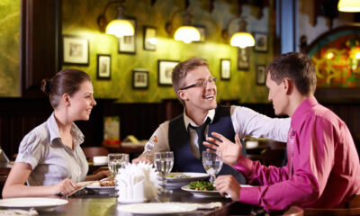 Três pessoas, uma mulher e dois homens estão à mesa de local público, conversando e sorrindo. À mesa, têm taças guardanapos e pratos