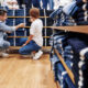 Homem de terno e mulher de calça jeans e camiseta se agacham para olhar prateleira de calças jeans em loja de jeans