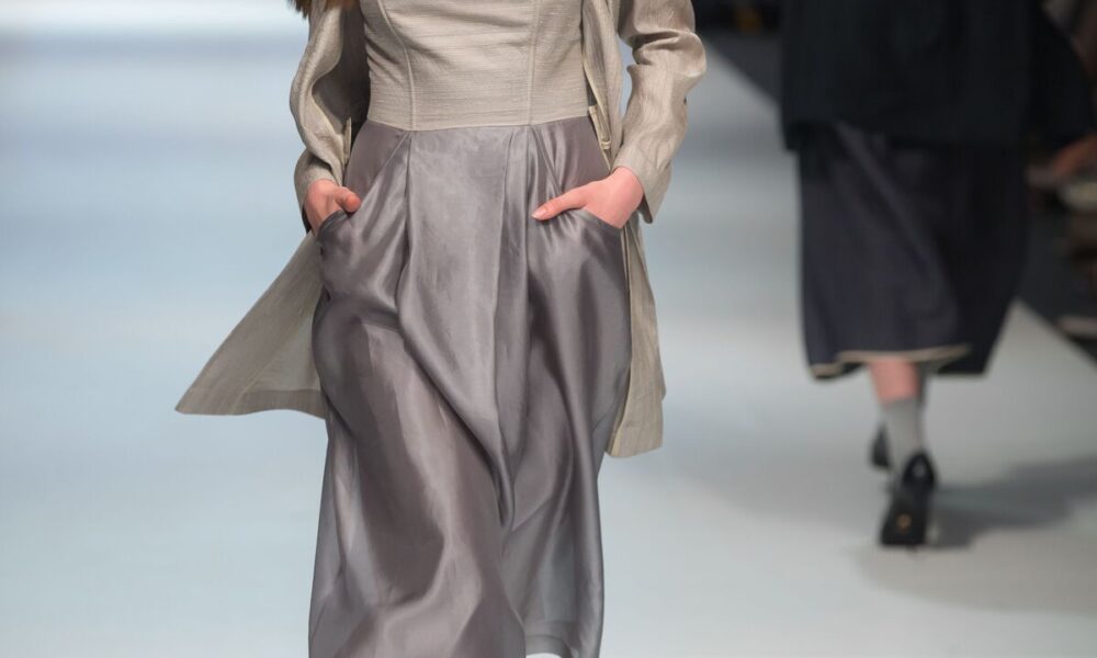 Modelo com rosto cortado da imagem desfila com mãos nos bolsos de vestido cinza-roxo-prateado, representando tendências da moda
