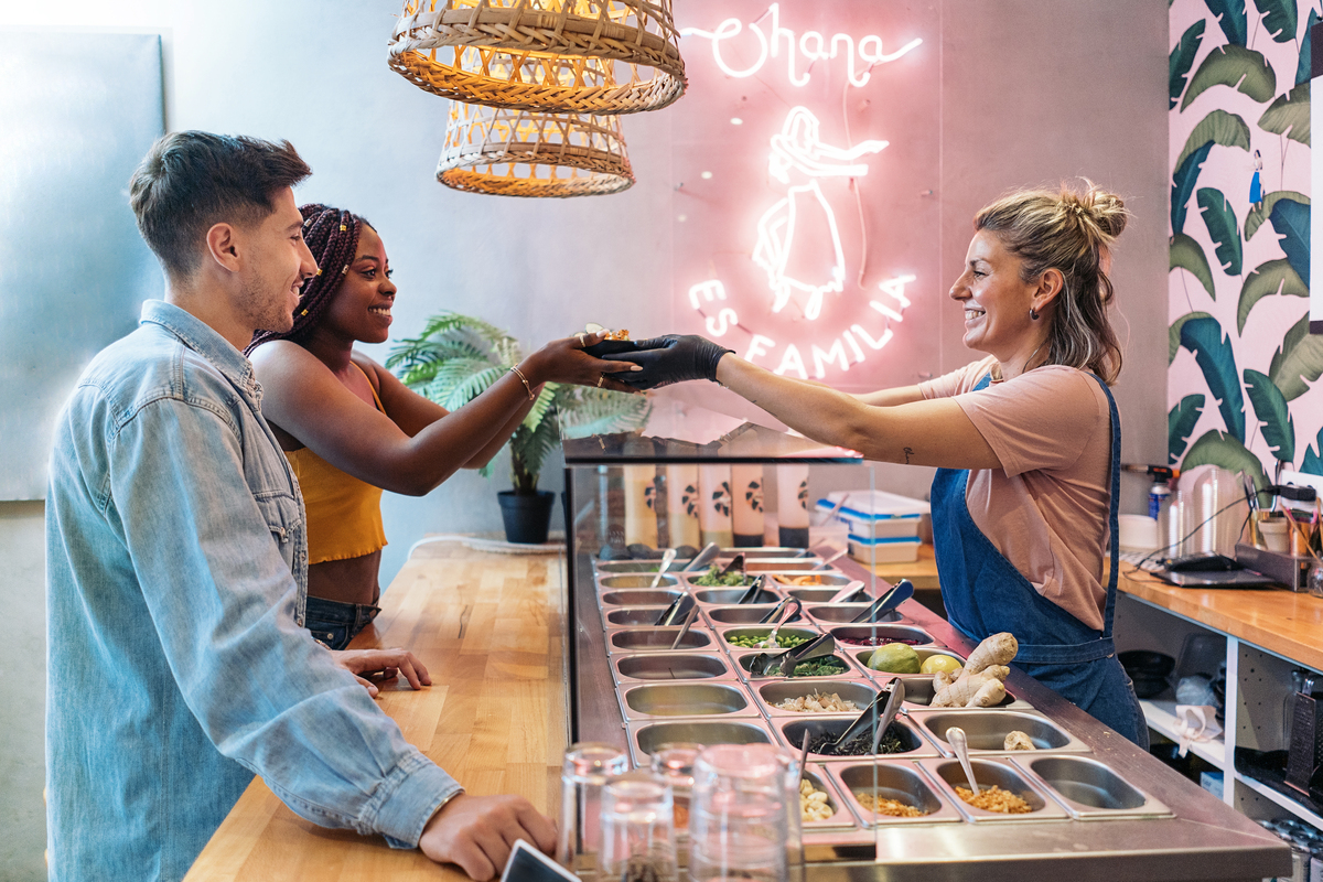 Um homem branco e uma mulher negra do lado de fora do balcão, enquanto vendedora loira entrega refeição em um estabelecimento colorido retratam experiência do cliente; experiência de compra