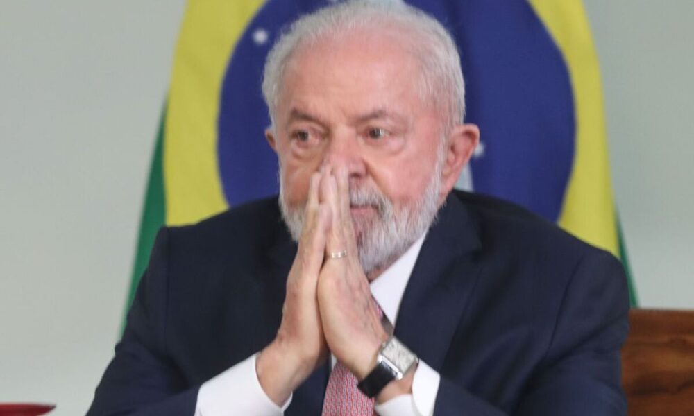 Lula de terno, com mãos em prece e expressão pessimista, com fundo da bandeira nacional; taxação compras internacionais