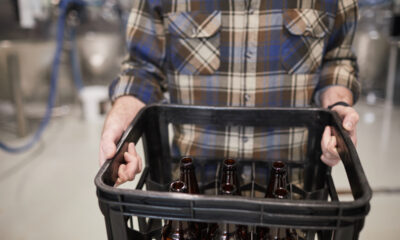 Homem com camisa xadrez escura carrega caixa preta de garrafas de cerveja vazias