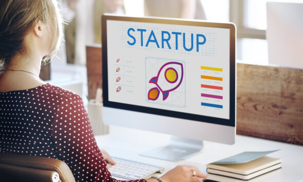 Mulher loira de cabelo amarrado está de costas para a câmera, sentada em uma mesa de frente a uma janela, mexendo em computador em que há o desenho de um foguete e a palavra "startup", representando empreendedorismo feminino