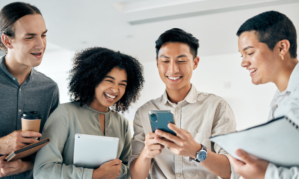 Homem oriental de camisa mostra celular para colegas, à esquerda, mulher com cabelo afro com caderno na mão, mais à esquerda, homem alto e branco com café na mão, e à direita, outro homem asiático com caderno na mão.