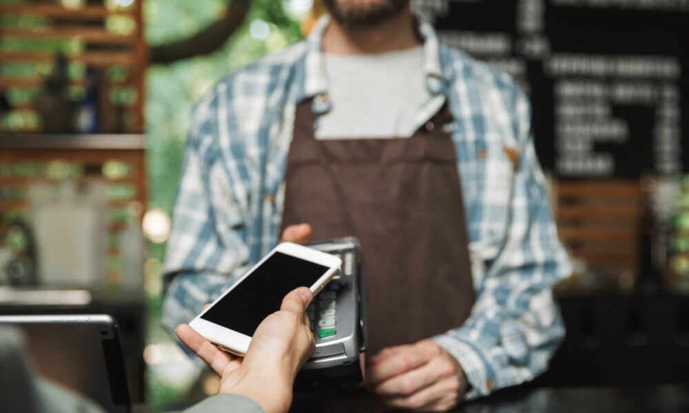 Homem barista com avental e camisa xadrez segunda máquina de cartão para cliente aproximar celular